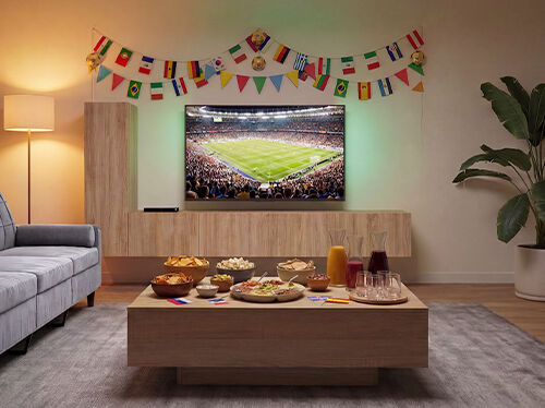 Sala de estar preparada para ver un partido de f&uacute;tbol, con un televisor que muestra un encuentro en un estadio, un sof&aacute; frente a una mesa de centro de madera llena de aperitivos y bebidas, y coloridas guirnaldas de banderas internacionales decorando la pared. Haz clic para ver los productos de la colecci&oacute;n.
