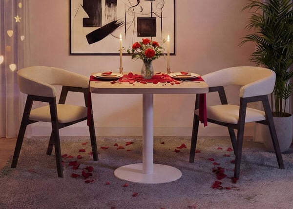 Escenario rom&aacute;ntico para cenar en un sal&oacute;n moderno con una peque&ntilde;a mesa redonda blanca y dos sillas tapizadas, dispuestas con platos, velas, un jarr&oacute;n con rosas rojas y p&eacute;talos de rosa esparcidos sobre la mesa y la alfombra, enmarcado por luces suaves en las cortinas, arte abstracto en la pared y una planta verde de interior para crear una acogedora atm&oacute;sfera de San Valent&iacute;n.