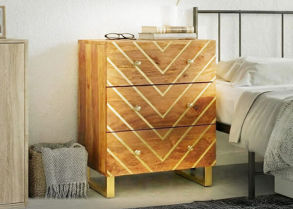 Mesilla de noche de madera con tres cajones y dise&ntilde;o geom&eacute;trico dorado, situada junto a una cama en un dormitorio luminoso.