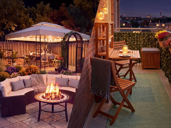 Espacios exteriores acogedores al atardecer, con un conjunto de jard&iacute;n con brasero y un cenador lleno de flores a un lado, y un balc&oacute;n decorado con muebles de madera, luces de farolillos y flores en maceta con vistas al perfil urbano al otro.