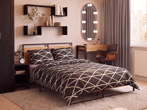 Dormitorio moderno con una cama de plataforma de metal negro y madera, vestida con ropa de cama en blanco y negro de dise&ntilde;o geom&eacute;trico, acompa&ntilde;ada por una mesita de noche a juego con un despertador de estilo vintage. Un armario alto de madera oscura se encuentra junto a estanter&iacute;as flotantes negras decoradas con libros, plantas y elementos decorativos. Frente a la cama, un tocador minimalista de madera con estructura negra se combina con una silla curva de nogal y un espejo ovalado con iluminaci&oacute;n integrada. Unas cortinas grises de suelo a techo enmarcan un gran ventanal que llena la estancia de c&aacute;lida luz natural, mientras que una suave alfombra color crema y un suelo de madera clara crean un ambiente acogedor y contempor&aacute;neo.
