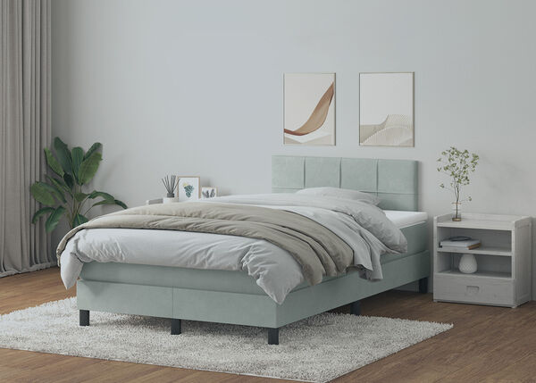 Cama boxspring en gris claro con cabecero panelado, ropa de cama blanca y mesita de noche y tocador de madera clara en un dormitorio decorado con estilo. 