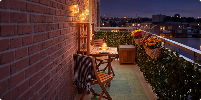  Balcón pequeño con sillas y mesa de madera, iluminado con farolillos y velas, decorado con flores en maceta y plantas, con vistas al perfil urbano de noche.
