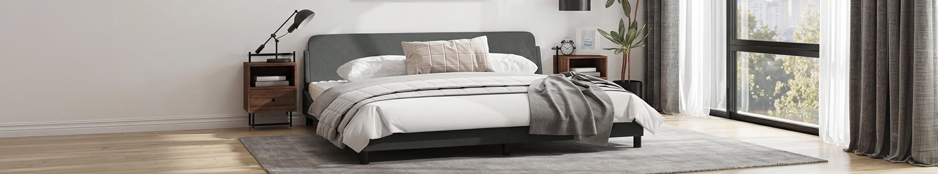 Plano general de un dormitorio moderno con una estructura de cama gris oscuro, ropa de cama blanca y un cojín beige con estampado de cuadrícula. La habitación cuenta con dos mesitas de noche de madera con lámparas, una planta alta en maceta y grandes ventanas con cortinas grises y blancas con vistas a árboles verdes.