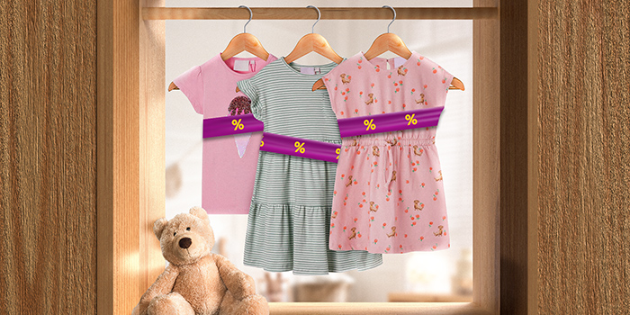 Ropa infantil colgada en perchas, envuelta en una banda promocional morada con iconos de descuento amarillos, colgada en una barra en interiores. Haz clic para ver los productos en oferta.