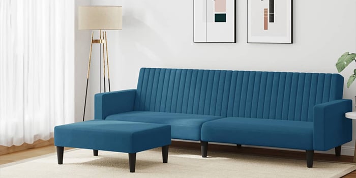 Sofá rinconera moderno de terciopelo azul, con tapizado acanalado vertical y un reposapiés a juego. El sofá tiene patas de madera negra y está situado en un salón luminoso, con cuadros enmarcados en la pared y una lámpara de pie en la esquina.